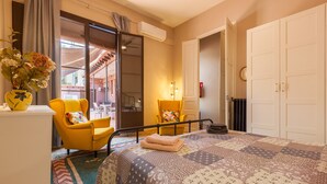 A hotel room with a bed, a yellow armchair, a small table with a vase of flowers, a radiator, and a door leading to a balcony.