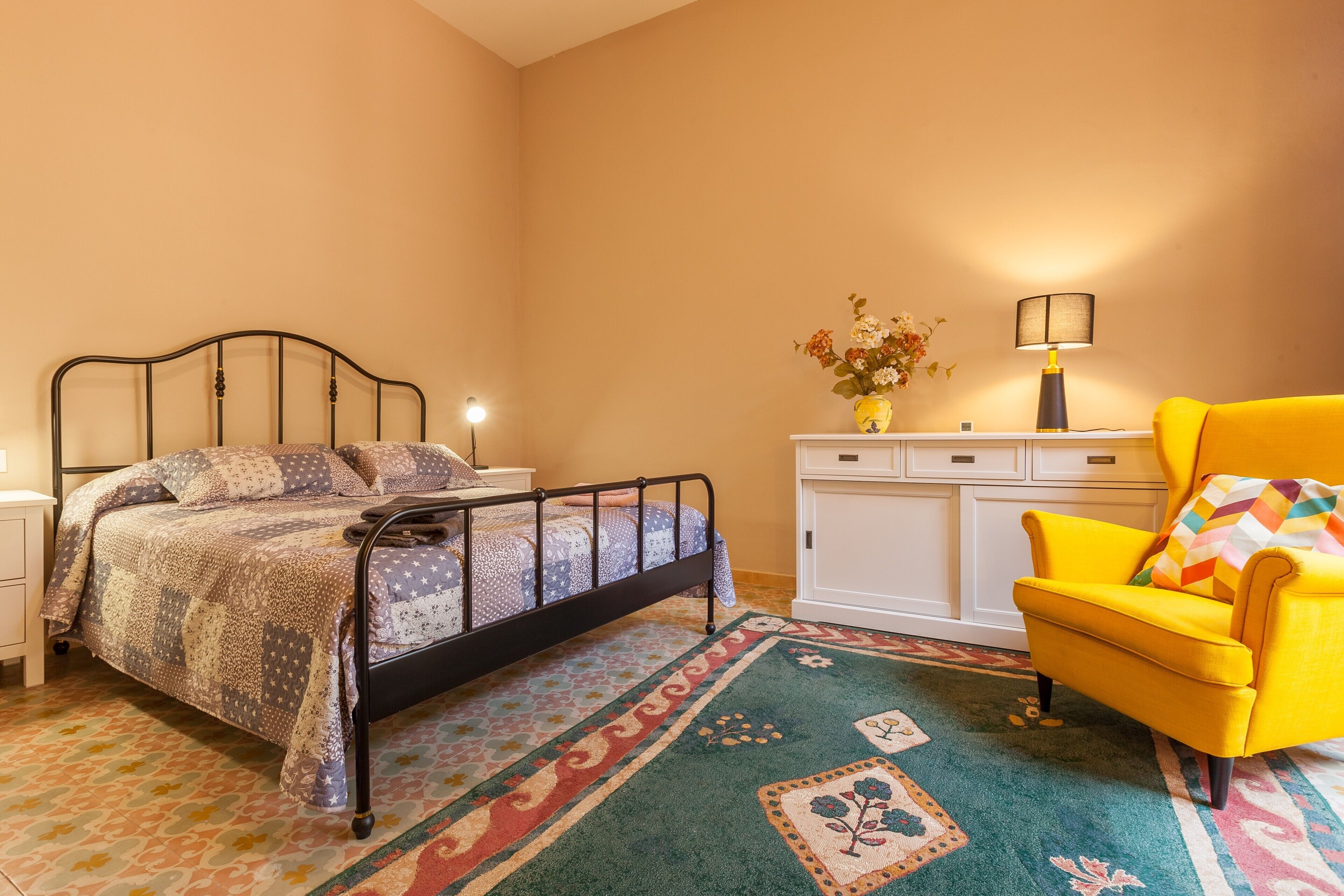 Un dormitorio con estructura de cama metálica, alfombra colorida, sillón amarillo, mesita de noche con lámpara y cómoda blanca.