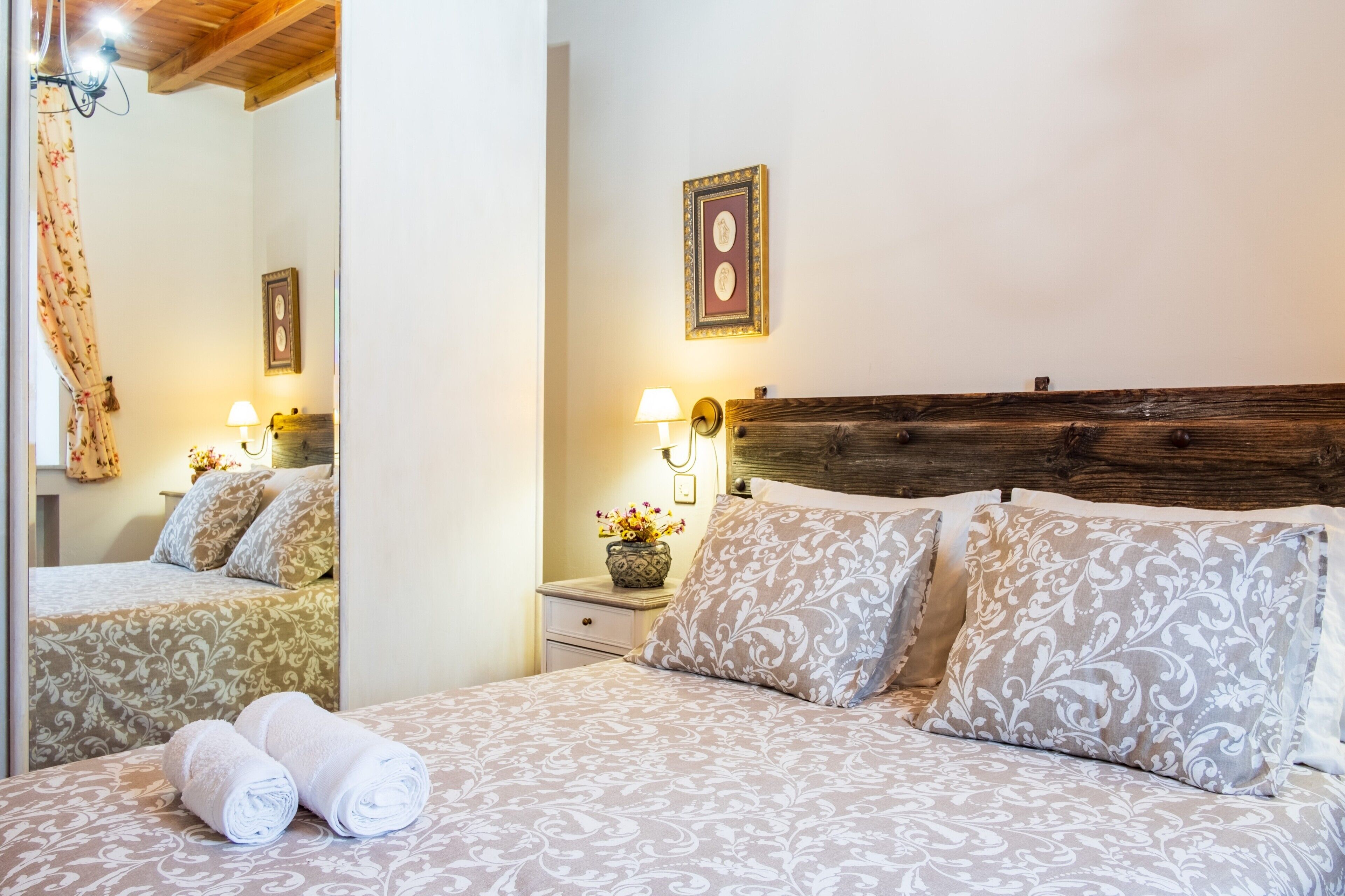 A neatly made bed with patterned bedding, a wooden headboard, bedside lamps, and a mirror reflecting the room.