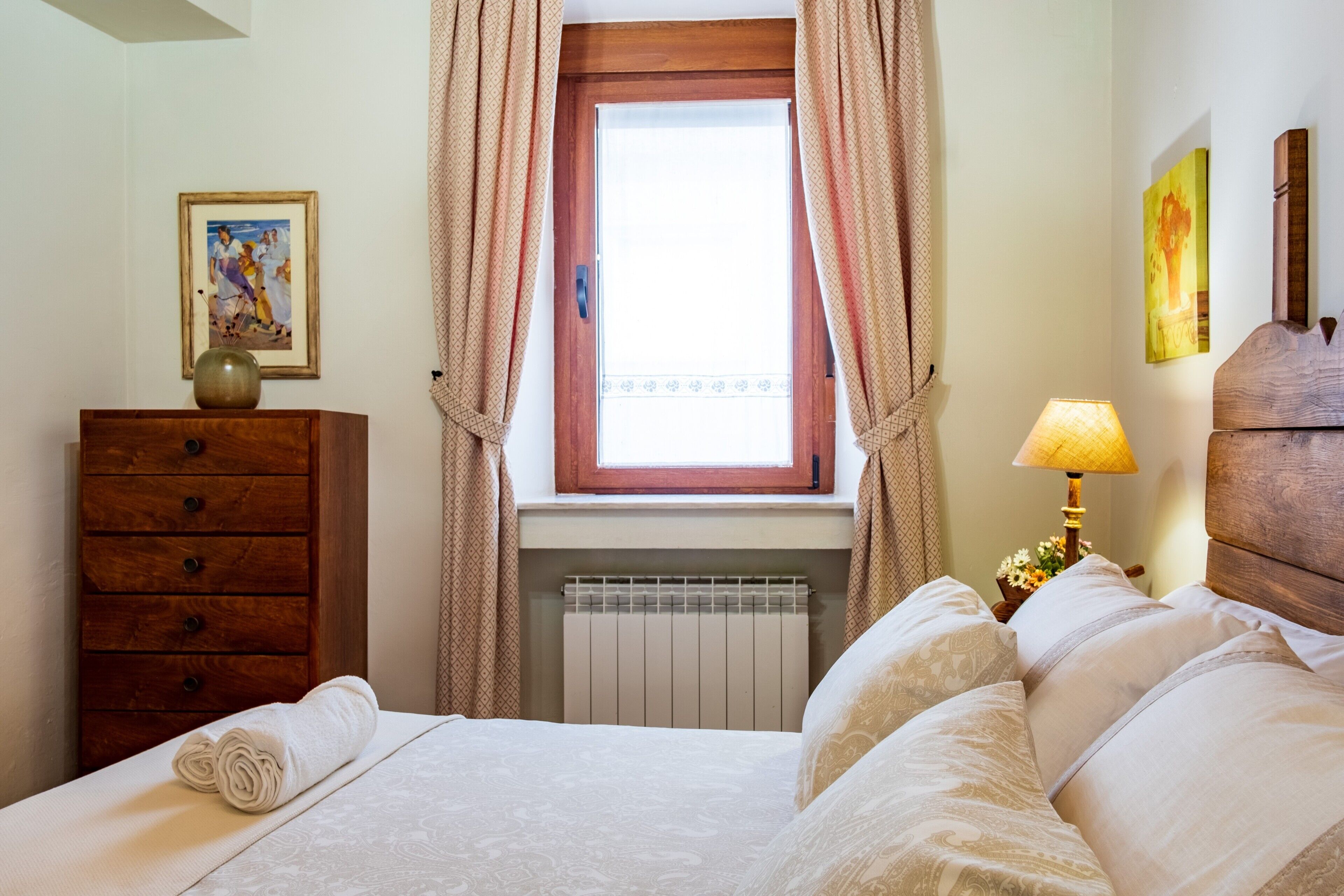 Un dormitorio con una cama, un aparador de madera, una ventana con cortinas, un radiador y una lámpara.