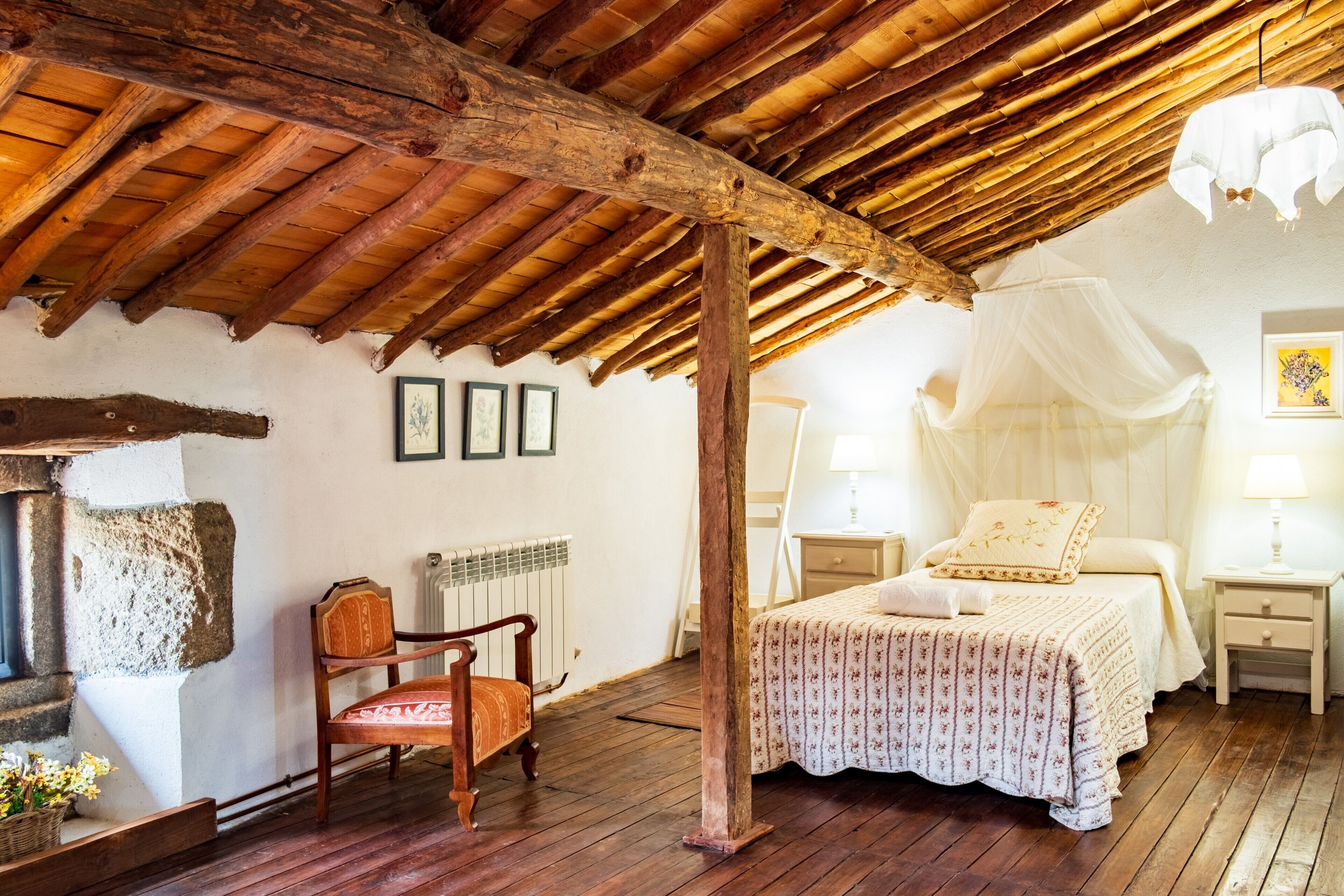 A rustic bedroom with a wooden ceiling, a bed with a canopy, a chair, and a bedside table with a lamp.