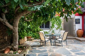 A stone patio with a table and chairs, surrounded by greenery and a tree.