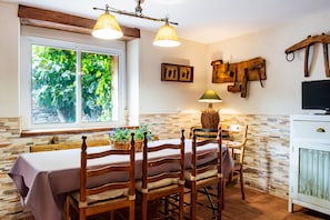 A dining area with a table set for four, wooden chairs, a chandelier, a television, and a window with greenery outside.