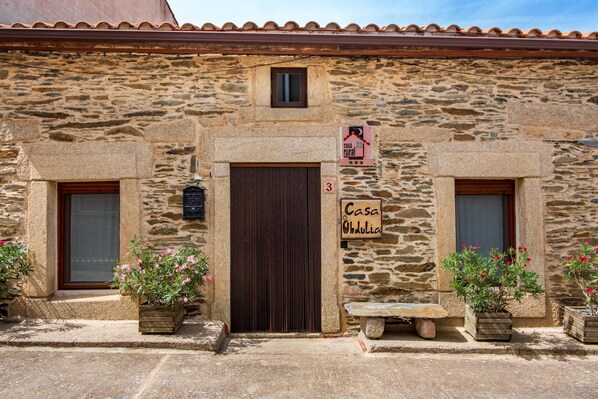 A rustic stone building with a central door, two side windows, and a small window above the door. There are potted plants on either side of the entrance and a bench in front.