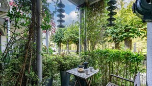A covered outdoor seating area with a table and bench, surrounded by greenery and a pergola with hanging lights.