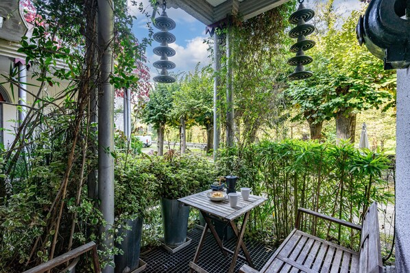 A covered outdoor seating area with a table and bench, surrounded by greenery and a pergola with hanging lights.