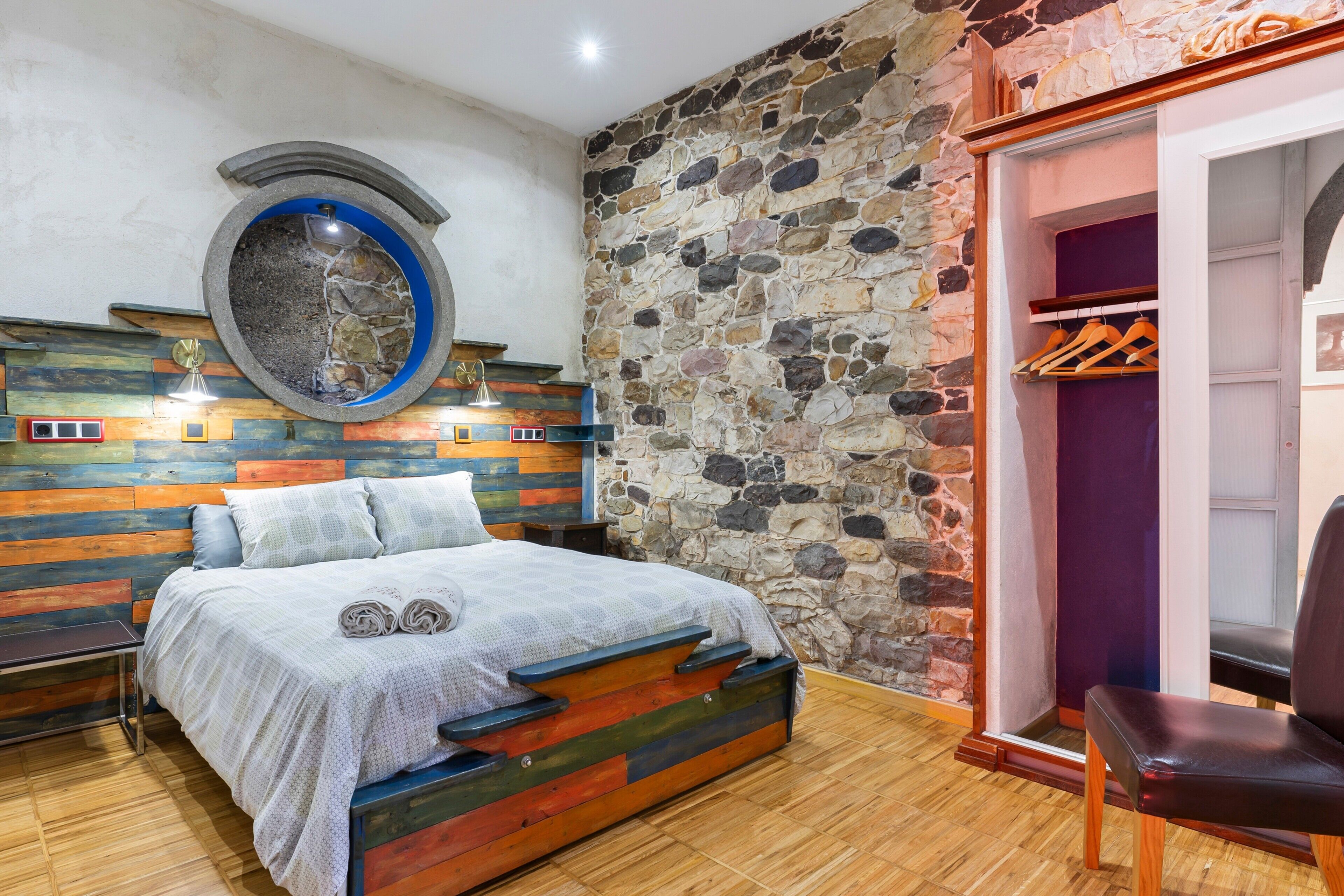 A bedroom with a stone wall, a bed with a colorful headboard, a circular window, and a wooden floor.