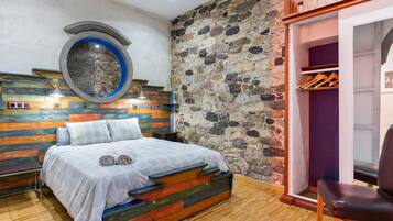 A bedroom with a stone wall, a bed with a colorful headboard, a circular window, and a wooden floor.