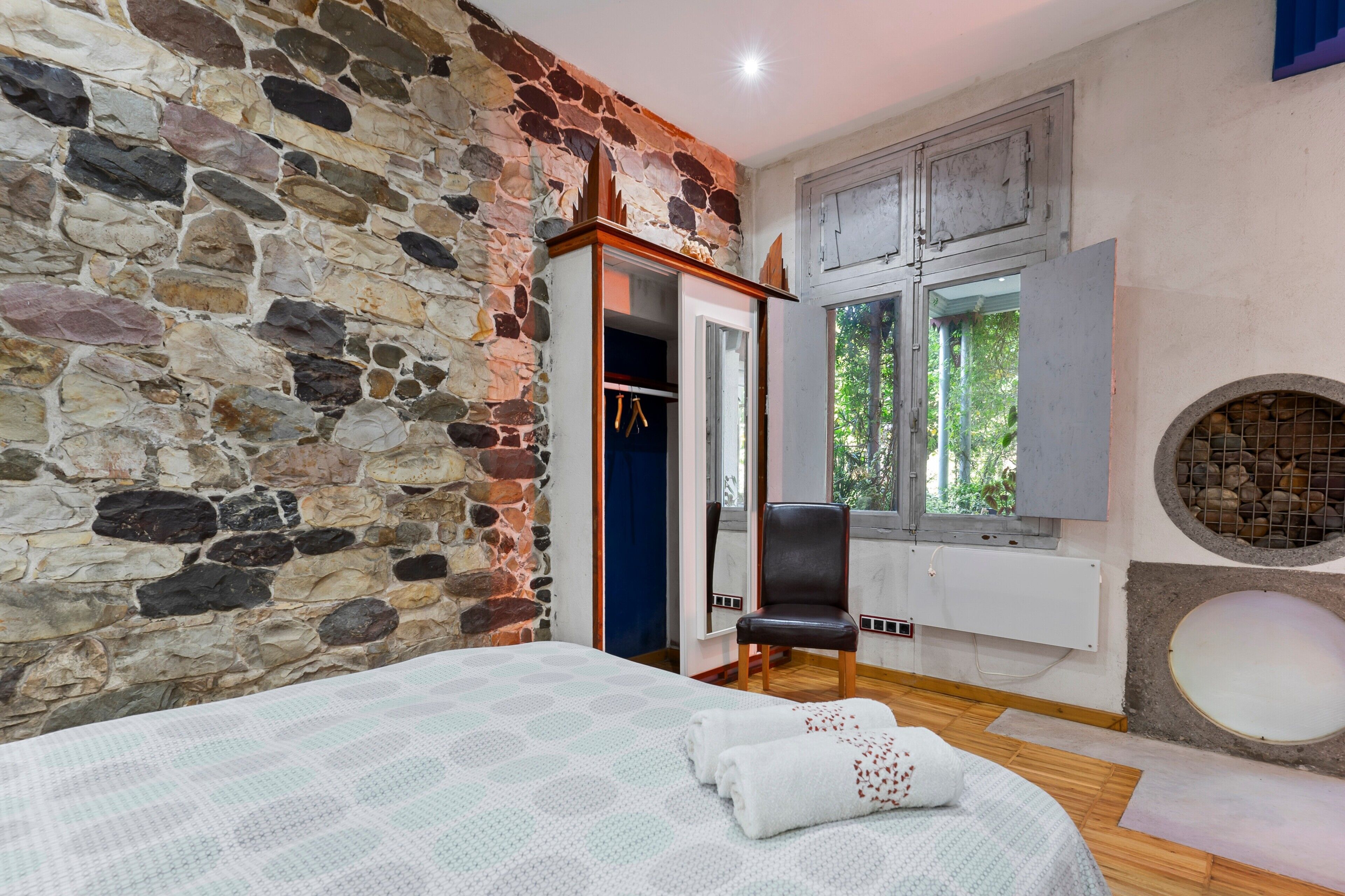 Una habitación con pared de piedra, una cama con edredón estampado, una silla, un armario y una ventana con vistas a vegetación.
