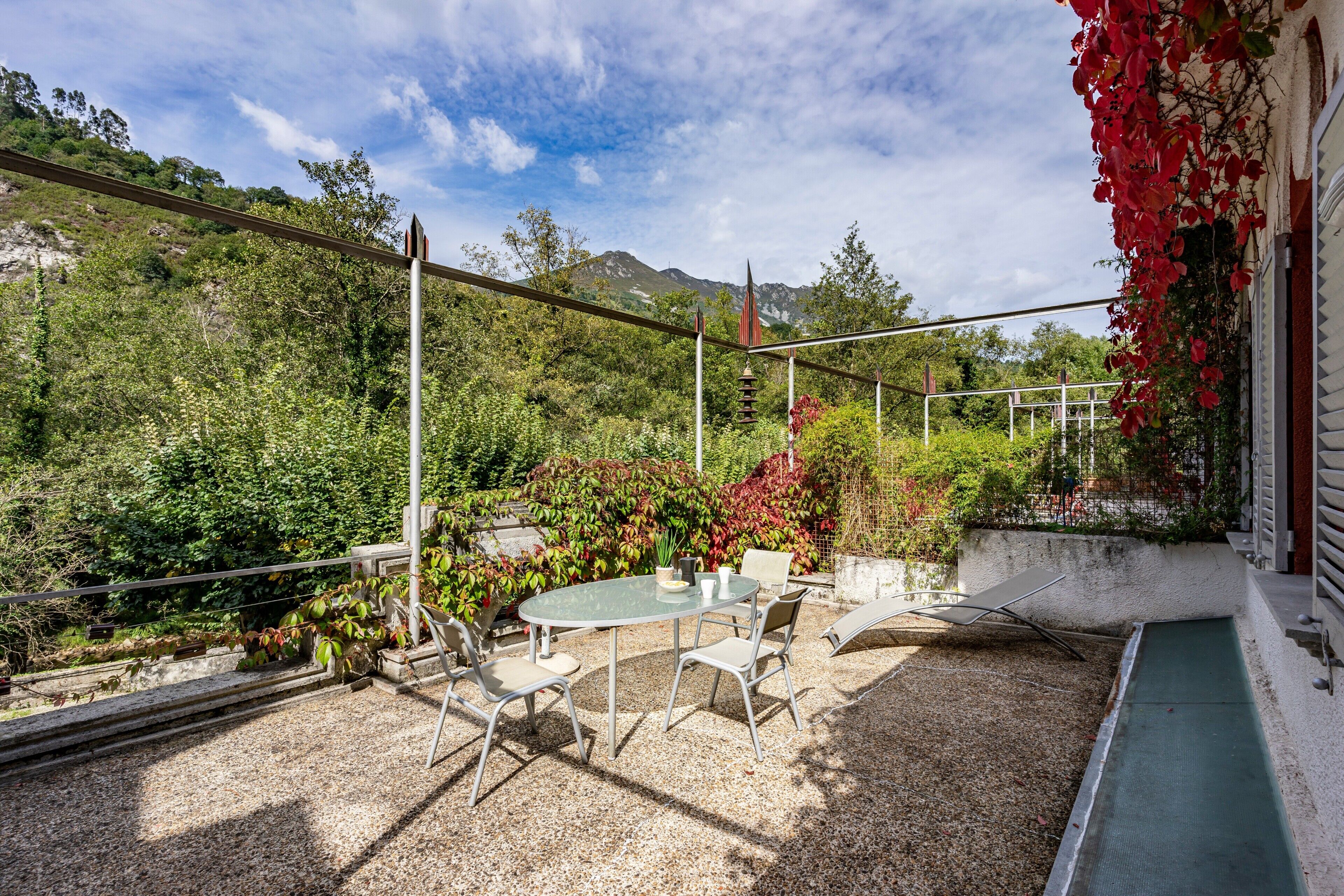 Een terras buiten met een tafel en stoelen, omgeven door groen en uitzicht op bergen.