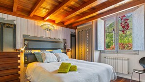 A bedroom with a wooden ceiling, a bed with white bedding, a nightstand, and a window with a view of greenery.
