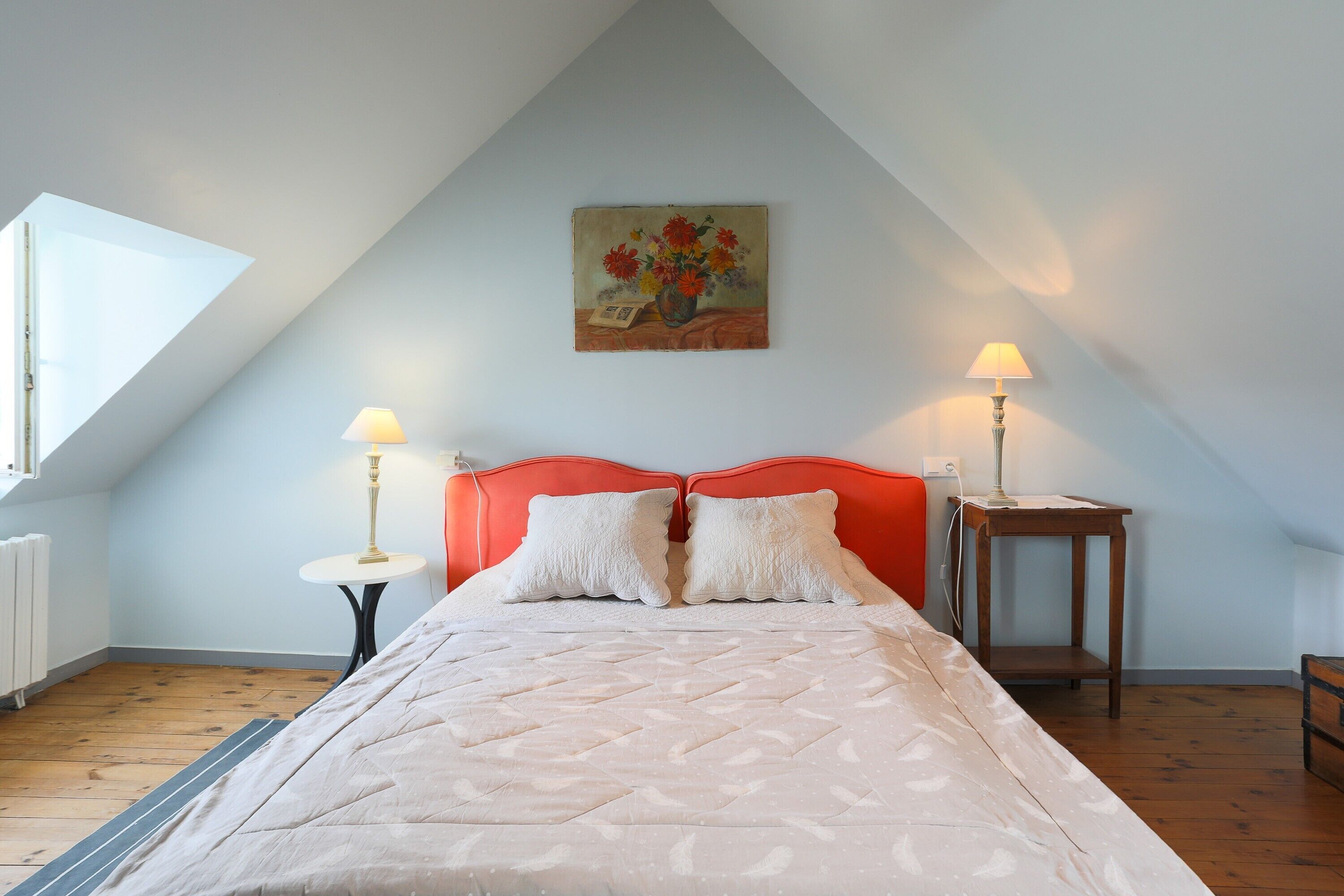 Une chambre à coucher avec un lit, une tête de lit rouge, deux tables de chevet avec des lampes, un tableau au mur et une fenêtre.