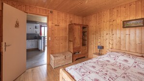 A wooden-paneled room with a bed, a wardrobe, a small table with a lamp, and a view into the kitchen. - Apartment 'La Charbonnière' with Mountain View, Private Garden and Wi-Fi (Cordon)