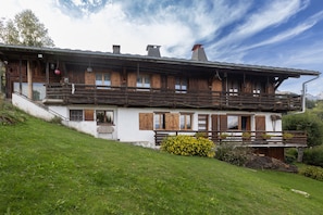 Een klassieke houten chalet met een balkon, een trap naar de ingang en een weelderige groene heuvel.