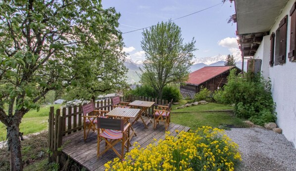 Una terraza de madera con mesas y sillas, rodeada de follaje y vistas a la montaña.