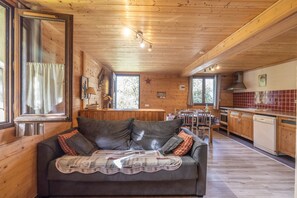 Un interior de cabaña acogedor, con techo de madera, un sofá cubierto con un edredón, una cocina con armarios de madera y un comedor.
