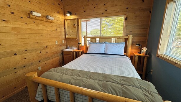 Un dormitorio de cabaña de madera con una cama, mesitas de noche, una ventana y un espejo.