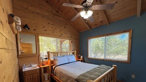 Un dormitorio de cabaña de madera con cama, tocador con espejo, ventilador de techo y ventanas con persianas.