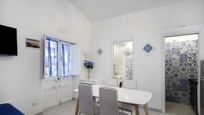 A compact living space with a dining area, a TV, a window with curtains, a mirror, and patterned walls. - Apartment 'Penisola Apartments' with Shared Terrace, Wi-Fi and Air Conditioning (Vico Equense)
