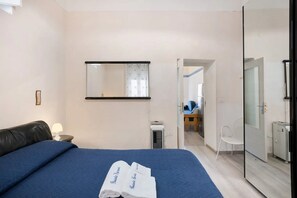 Habitación de hotel con cama, mesita de noche, espejo, calentador de pared, silla y puerta que da a un baño.