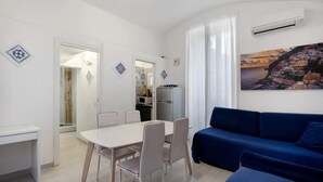 A modern living room with a blue sofa, a dining table with chairs, a kitchen area, and a painting of a coastal scene. - Apartment 'Penisola Apartments' with Shared Terrace, Wi-Fi and Air Conditioning (Vico Equense)