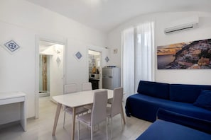 Un salon moderne avec un canapé bleu, une table à manger avec des chaises, un coin cuisine et un tableau représentant un paysage côtier.