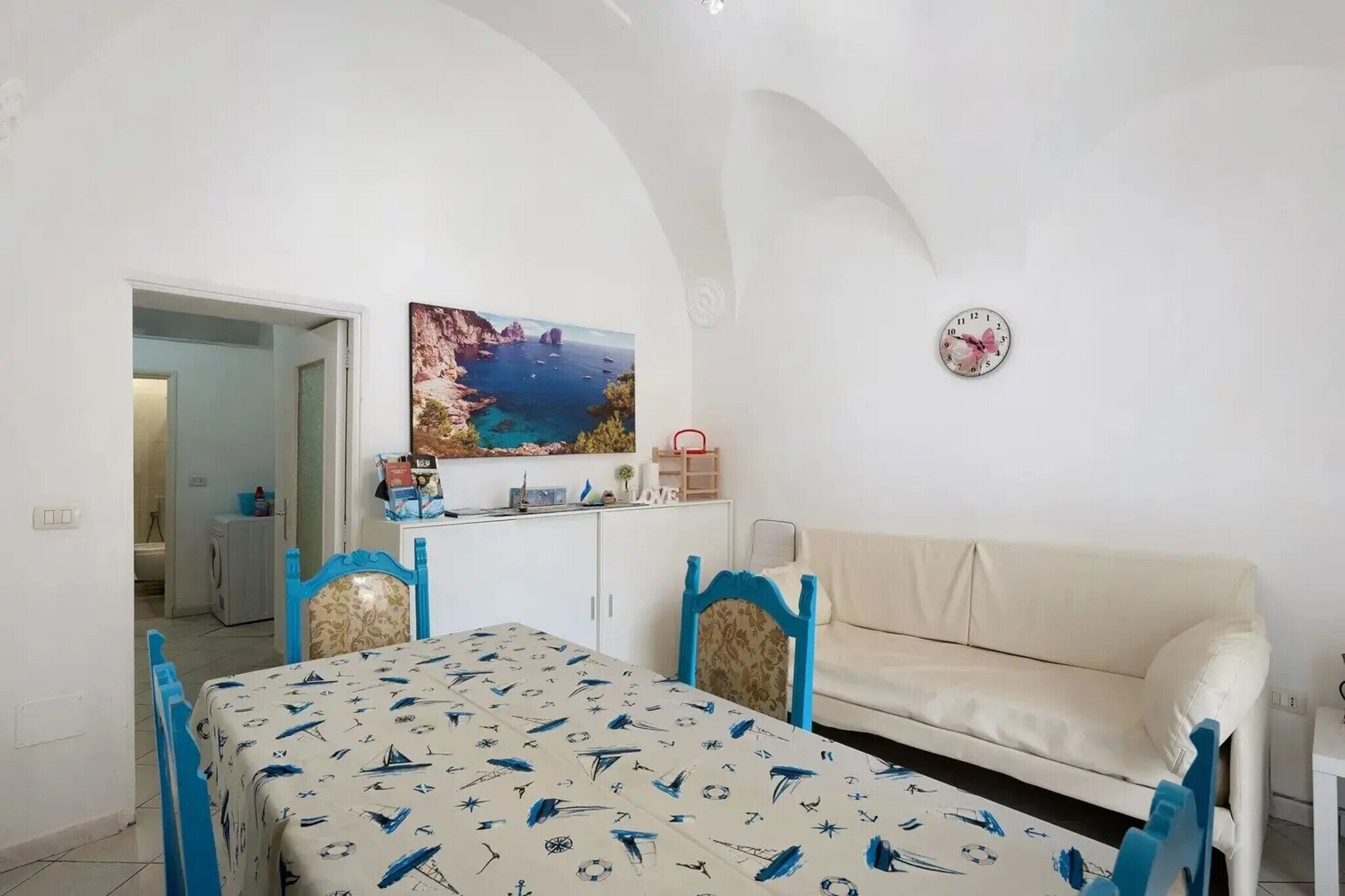 A room with a dining table, chairs, a sofa, a painting, a clock, and a view of the sea.