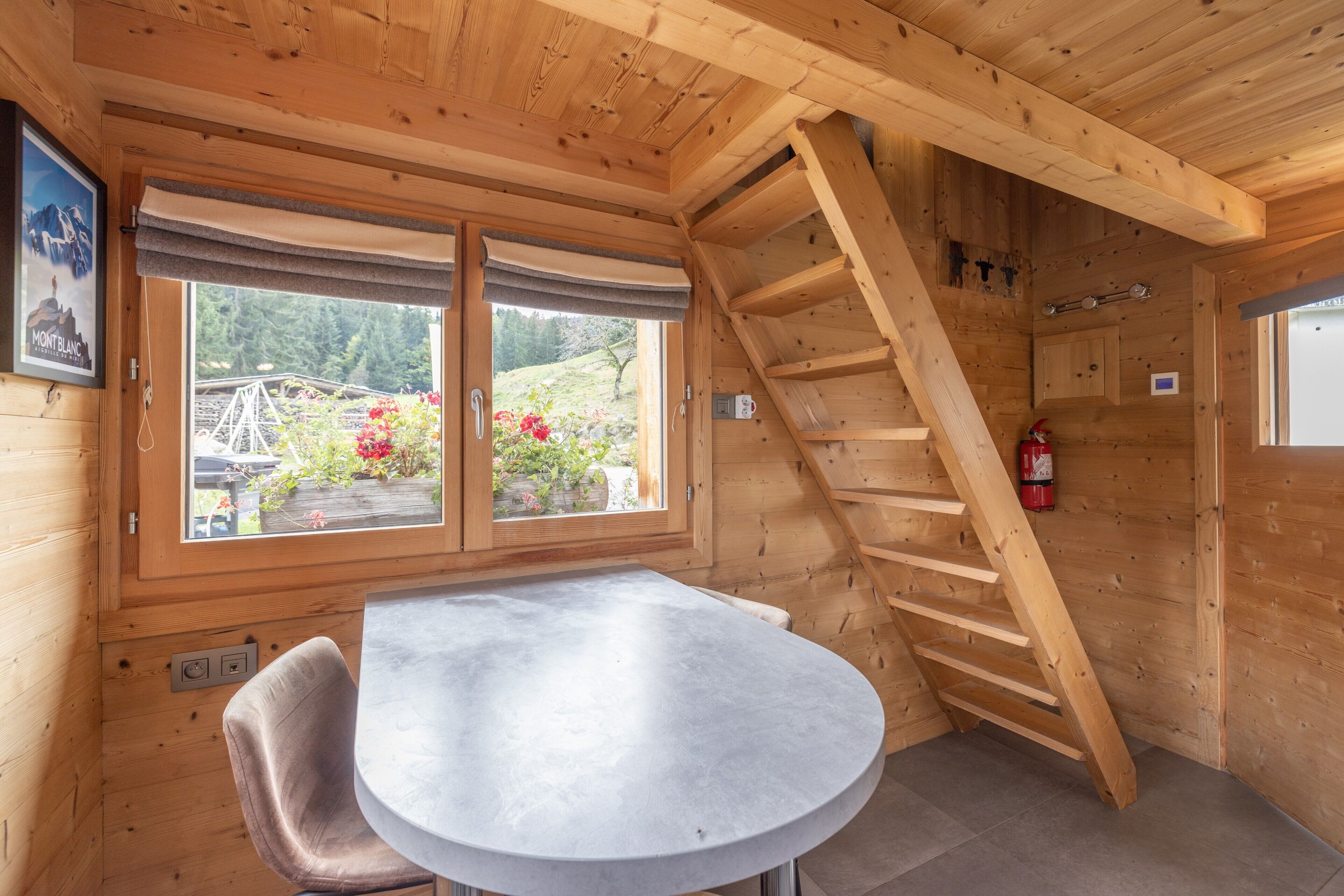 Interior de madeira com mesa, cadeiras, janela com vista para vegetação e escada de madeira.