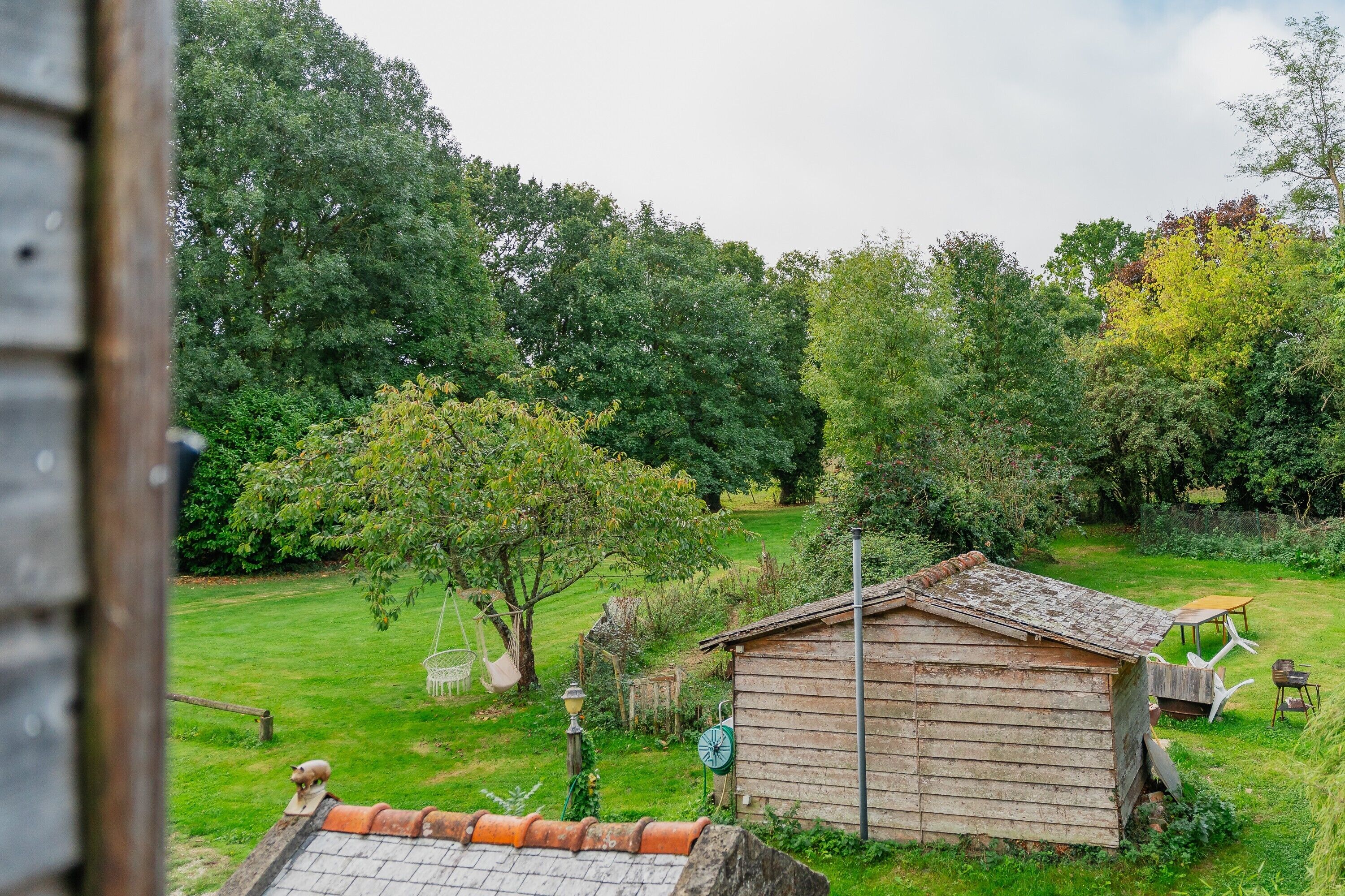 A rustic wooden shed in a lush garden with a lawn, a hanging chair, and a small table.