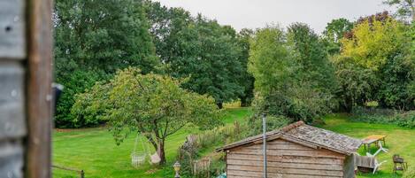 A rustic wooden shed in a lush garden with a lawn, a hanging chair, and a small table.