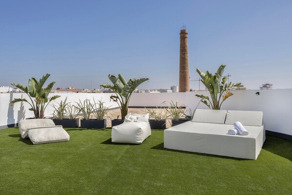 Eine Dachterrasse mit einer weißen Couch, zwei Sitzsäcken und Topfpflanzen. Im Hintergrund ist ein hoher Schornstein zu sehen.