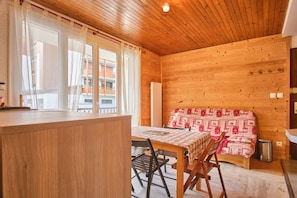 A wooden cabin interior with a dining table, chairs, a bench with a patterned cover, and a window with curtains. - Apartment 'Shrischss Joder' with Balcony (Les Deux-Alpes)