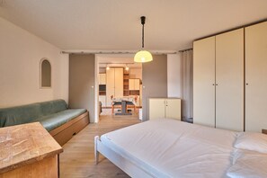A bedroom with a bed, a green sofa, a wooden table, a wardrobe, and a kitchen area in the background.