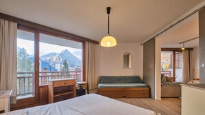 A room with a bed, a desk, a chair, a sofa, and a view of mountains. - Apartment 'Meijotel' with Mountain View and Balcony (Les Deux-Alpes)