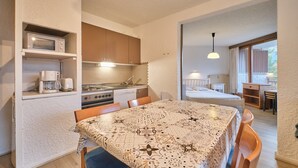A compact kitchen with a microwave, coffee maker, and stove, a dining area with a patterned tablecloth, and a bedroom with a bed and window. - Apartment 'Meijotel' with Mountain View and Balcony (Les Deux-Alpes)