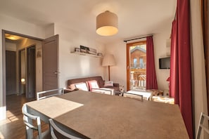Una stanza con un tavolo da pranzo, sedie, un divano, una lampada, una TV e una porta in legno che dà su un'area esterna.