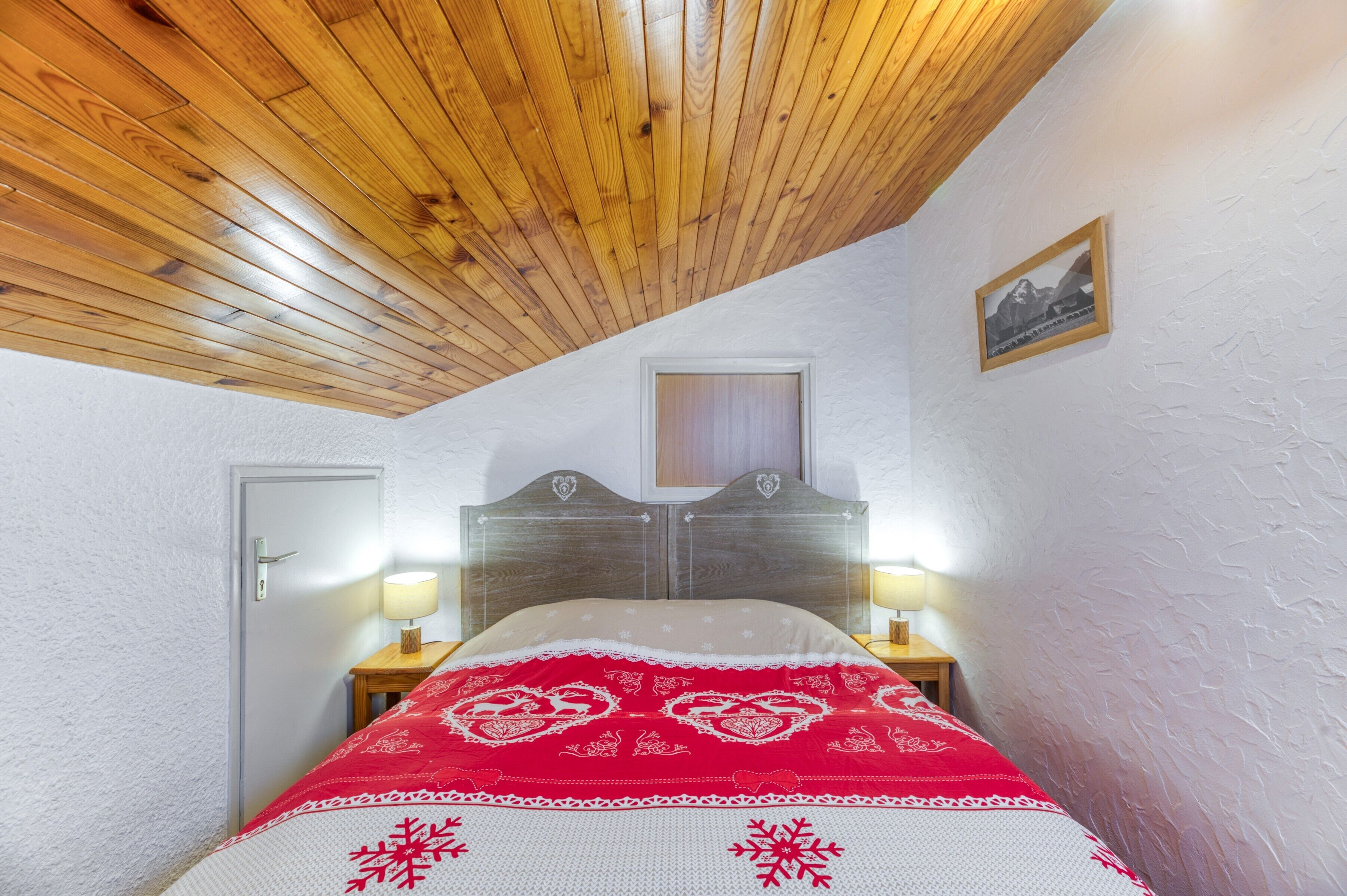 Ett sovrum med ett träpanelat tak, en säng med ett rött och vitt sängöverkast, två sänglampor, en dörr och en inramad tavla på väggen.