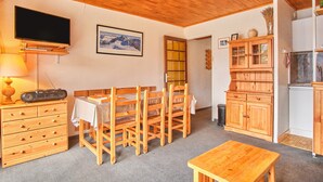 A room with a wooden ceiling, a television, a dining table with chairs, a cabinet, and a kitchen area.