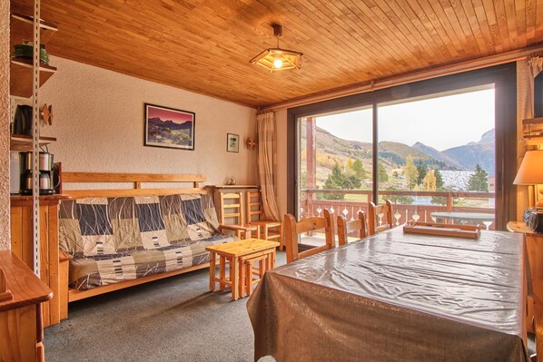 A wooden interior with a sofa, dining table, chairs, a view of mountains, and a painting on the wall.