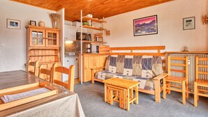 A cozy room with a wooden ceiling, a sofa with a quilted cover, a dining table with chairs, a wooden cabinet, and a radiator.