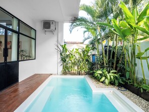 A modern indoor pool with a wooden deck, surrounded by lush tropical plants and a white wall with a window and air conditioning unit. - Berawa Factory Lofts (Canggu)