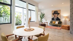 A modern living room with a round dining table, comfortable chairs, a sofa, and a large window with curtains. - Berawa Factory Lofts (Canggu)