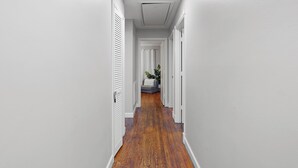 Un couloir étroit aux murs blancs et au plancher en bois, qui mène à un espace détente agrémenté d’une plante.