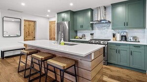 Eine moderne Küche mit grünen Schränken, einem Edelstahlkühlschrank, einem Gasherd und einem eingebauten Backofen. Zwei Barhocker und eine Bank befinden sich in der Nähe der Kücheninsel.