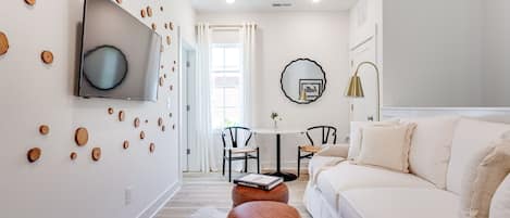 A modern living room with a white sofa, a brown ottoman, a flat-screen TV mounted on the wall, and a dining area with a table and chairs.