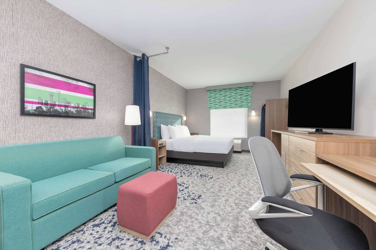 A hotel room with a teal sofa, a red ottoman, a bed with white linens, a desk with a flat-screen TV, and a patterned carpet.