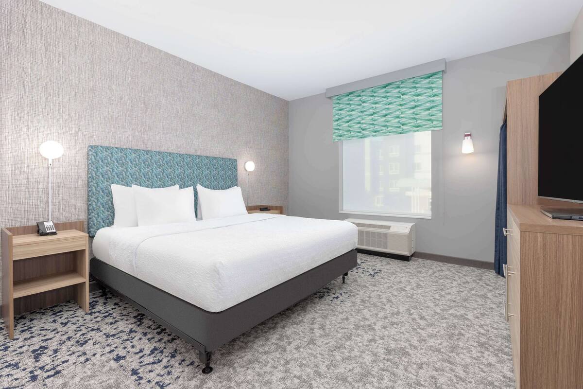 A hotel room with a bed, bedside tables, a TV, a window with blinds, and a wall-mounted heater.