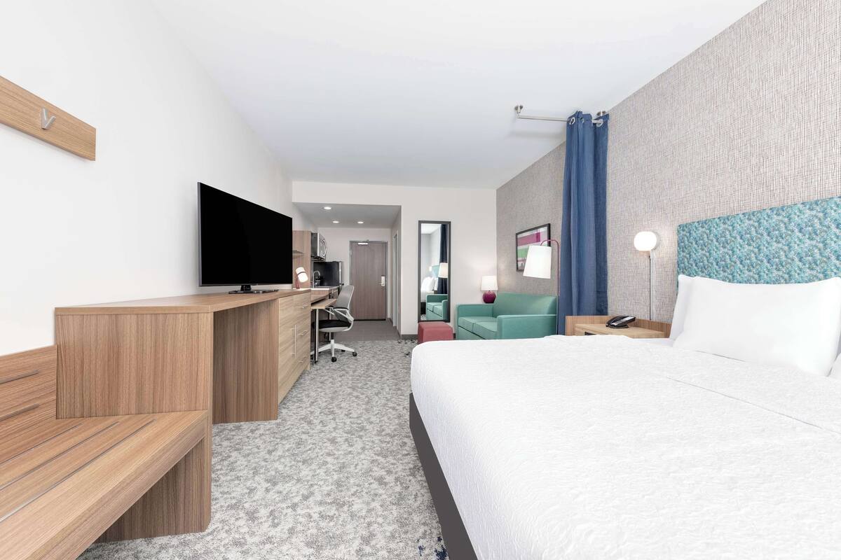 A hotel room with a large bed, a desk with a flat-screen TV, a chair, a nightstand, and a lamp.