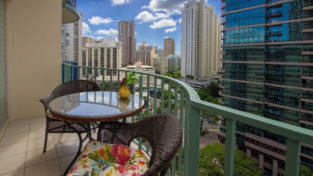 A balcony with wicker furniture, a glass table, and a view of a cityscape with tall buildings. - Cozy two bedroom, sleeps 6. Wifi, full kitchen, balcony, and pool. (Honolulu)