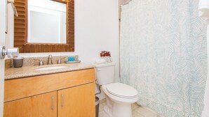 A bathroom with a wooden vanity, a white toilet, a sink with a mirror, and a shower curtain with a leaf pattern. - Cozy two bedroom, sleeps 6. Wifi, full kitchen, balcony, and pool. (Honolulu)
